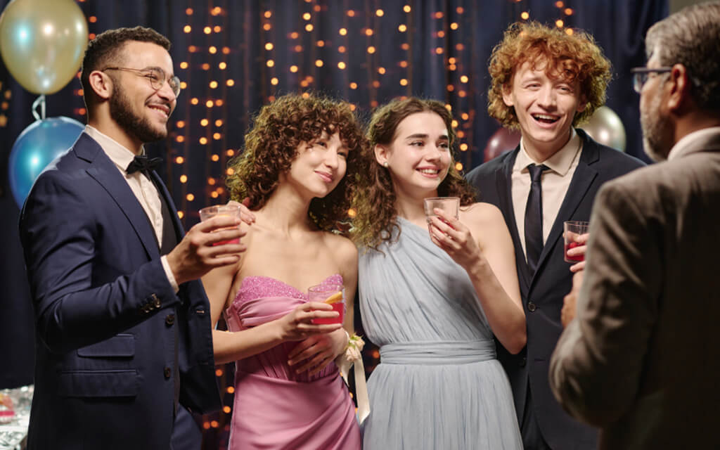 Group of younger people standing with drinks in their hands while smiling and talking to an older gentleman